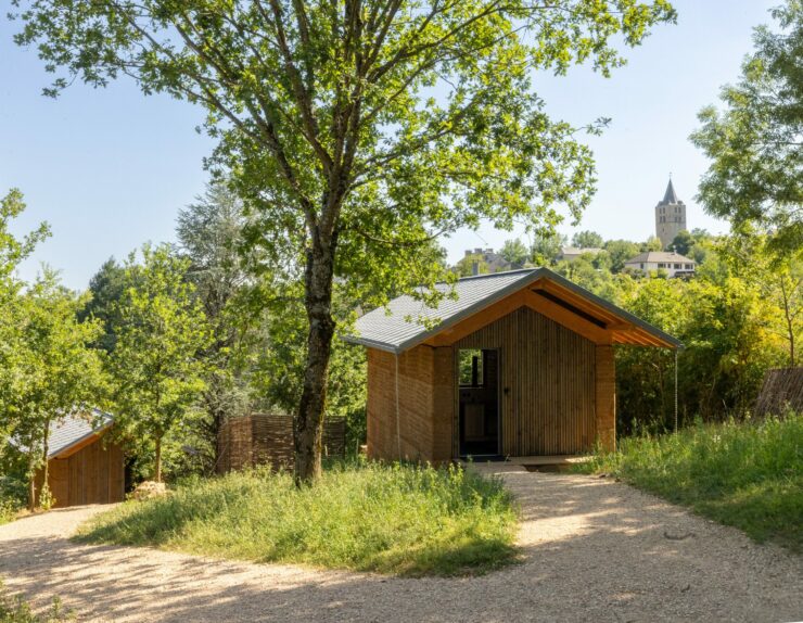 Lodges en pisé et bois nichés dans la verdure, avec le clocher de Sauveterre-de-Rouergue en arrière-plan – Les Safranières